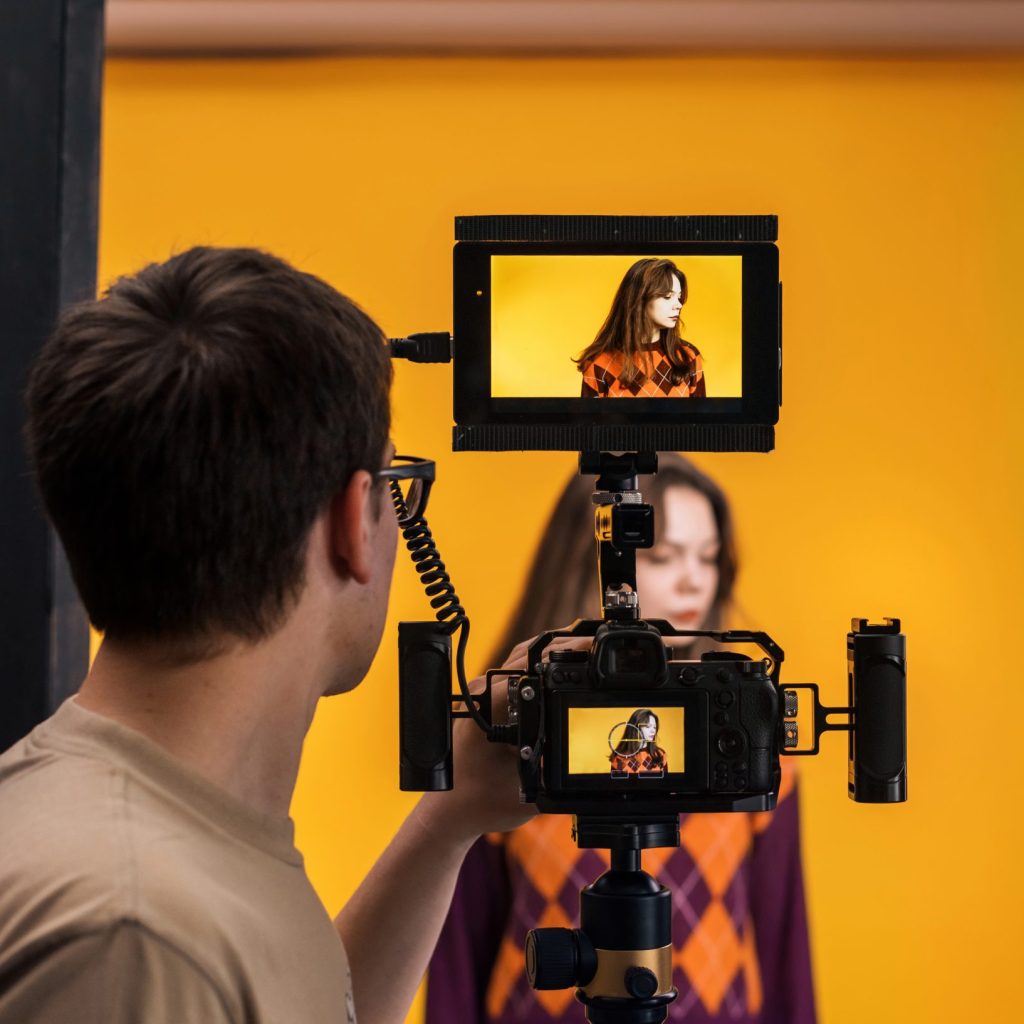 View of a professional photographer shooting a young woman using a camera with external display, yellow background